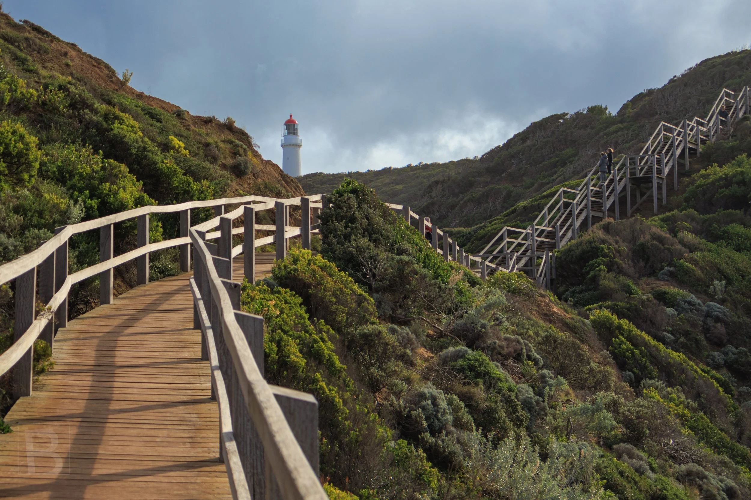 A weathered boardwalk leads through windswept vegetation toward a
lighthouse, connecting practical coastal access with navigation and
shelter.