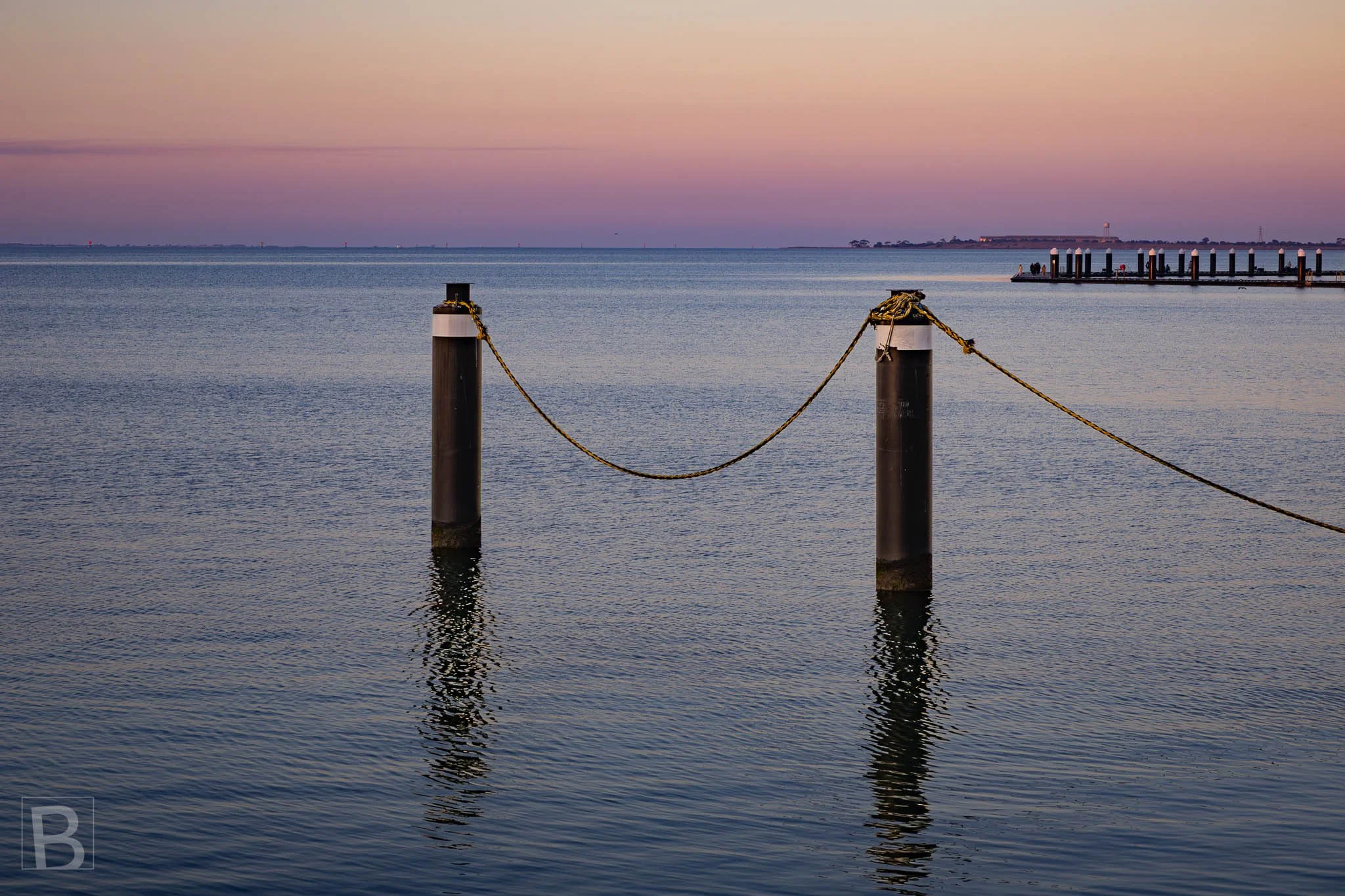 A simple chain suspended between posts draws a quiet line across
reflective water, with distant piers fading into the evening.