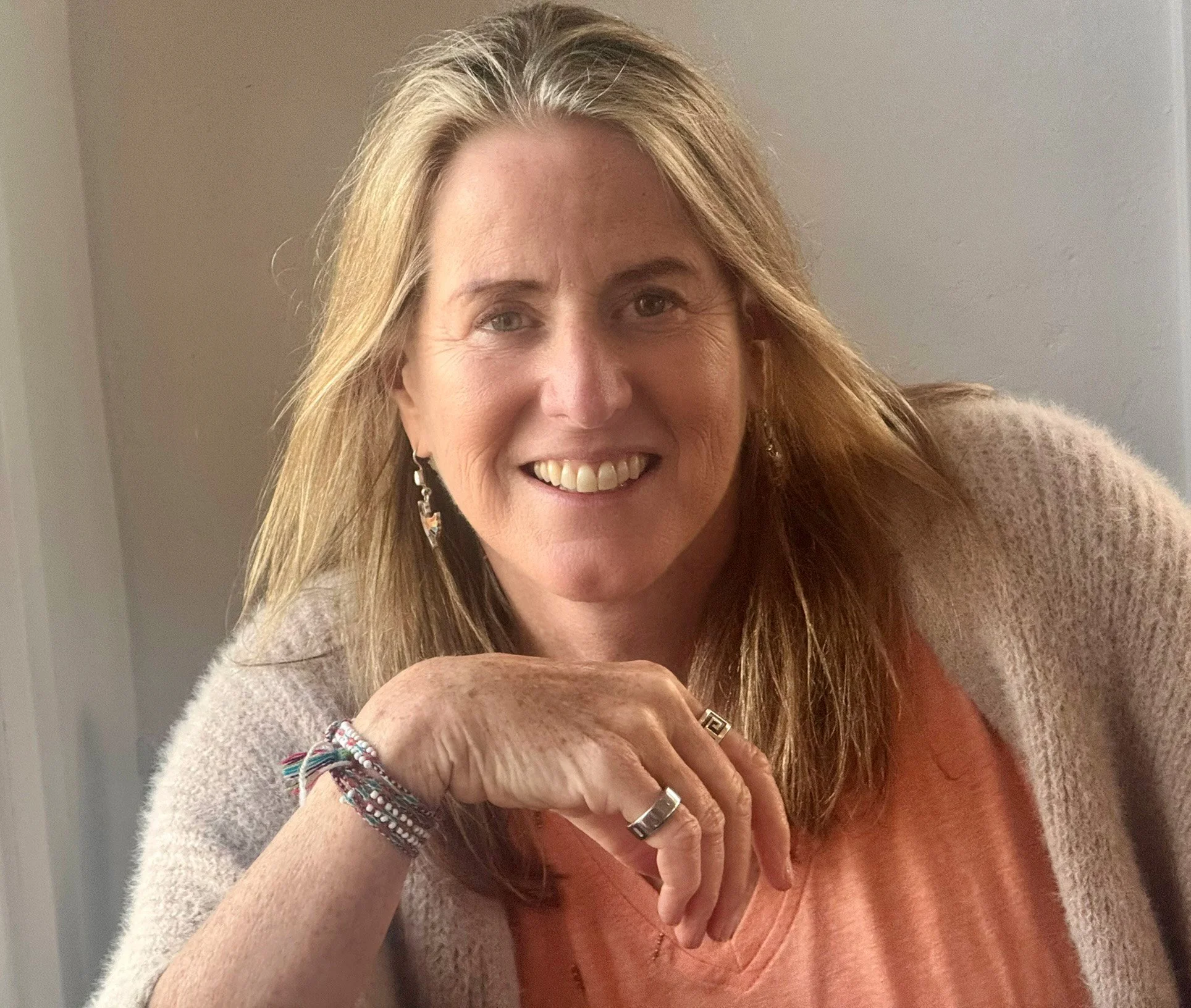 A woman with long blonde hair and light skin smiling, wearing earrings, rings, a bracelet, a peach-colored shirt, and a beige sweater, resting her chin on her hand.