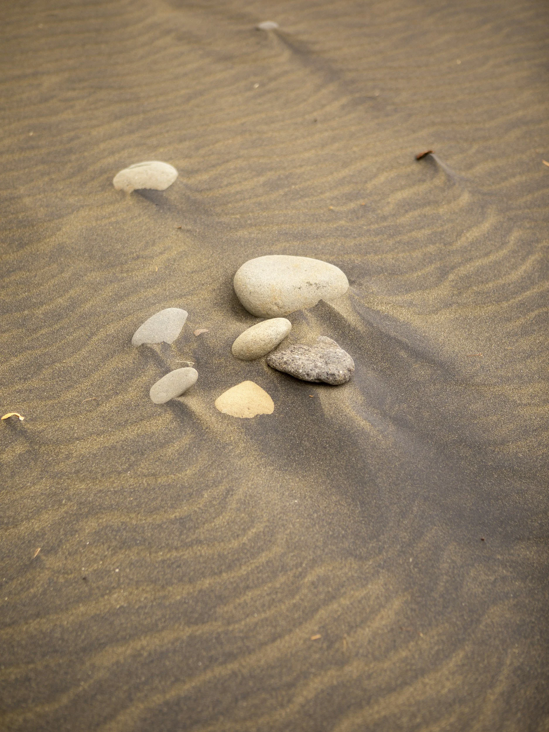 A sandy beach with several scattered smooth stones and pebbles, with some gentle ripples in the sand.