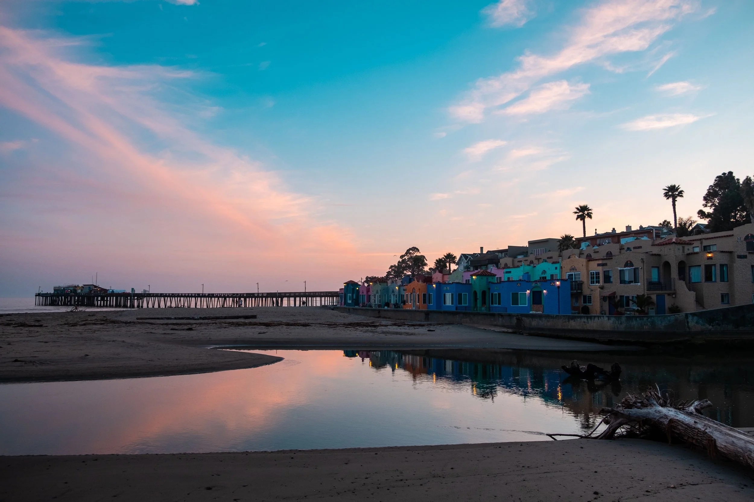 Colorful houses along a beach during sunset with a pier extending into the water, palm trees, and a sky with pink and blue clouds.