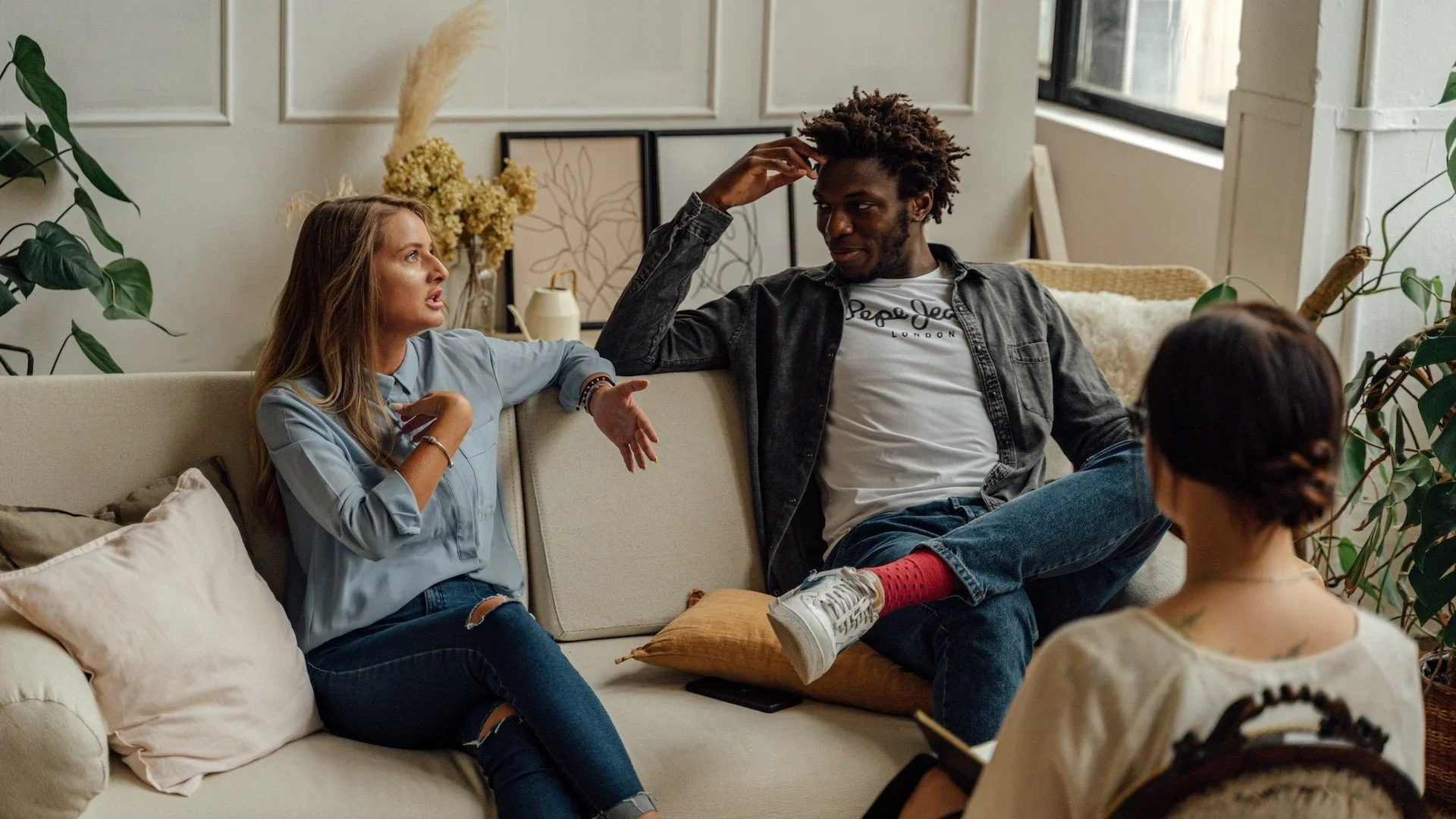 Four people sitting on a beige sofa in a living room having a conversation. There are decorative plants, artwork, and a window in the background.