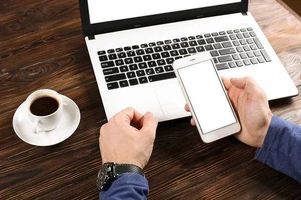 Person holding a smartphone near a laptop on a wooden desk with a cup of coffee.