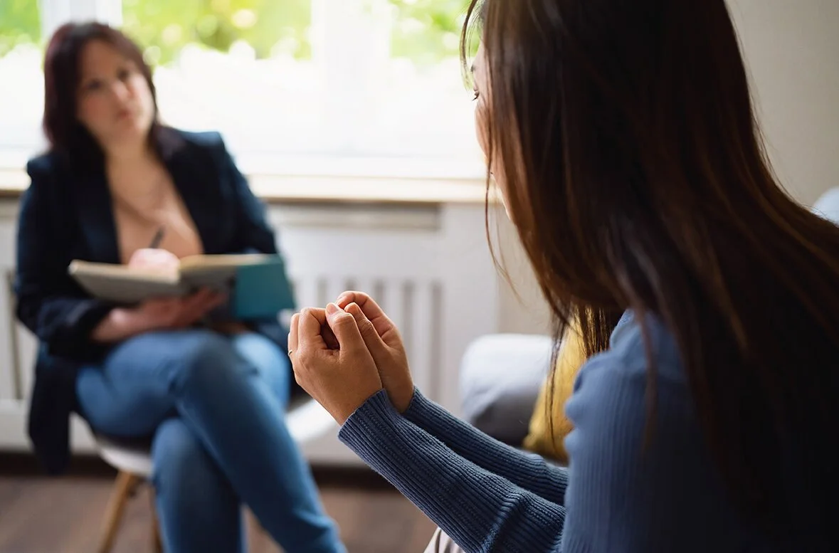 A woman with long dark hair, wearing a blue sweater, praying with clasped hands, during therapy session. In the background, another woman with red hair, in a dark jacket and jeans, sitting on a chair, holding a notebook.