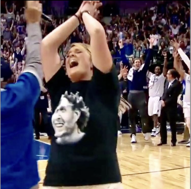 A woman celebrates excitedly on a basketball court surrounded by others who are cheering and raising their hands in celebration.