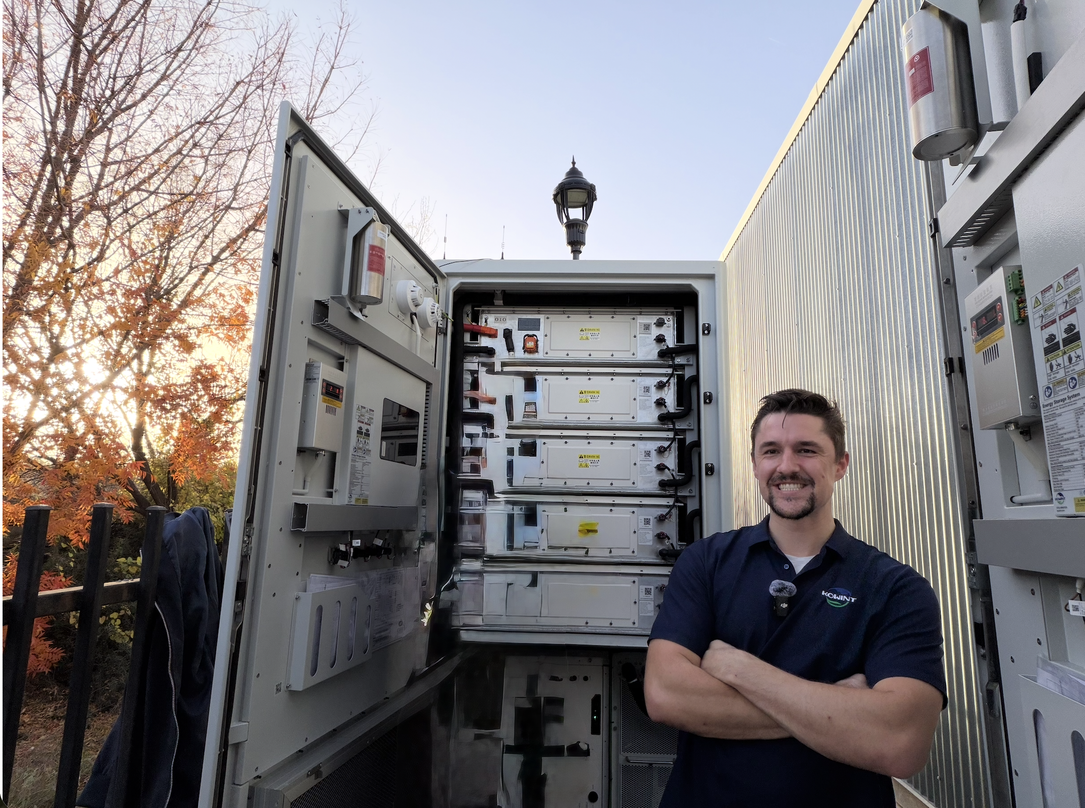 A man with short brown hair and a beard smiling while standing in front of an outdoor electrical equipment setup, with trees with autumn-colored leaves in the background.