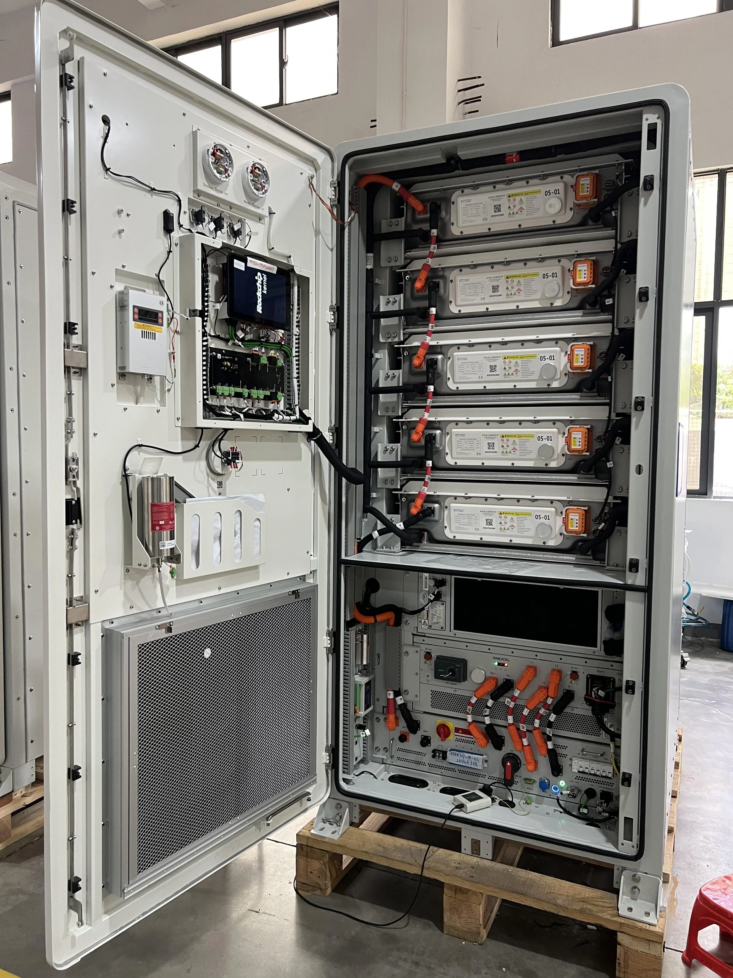 An open electrical control cabinet with various electronic components, wiring, cooling fans, and a small display screen inside a factory or industrial setting.