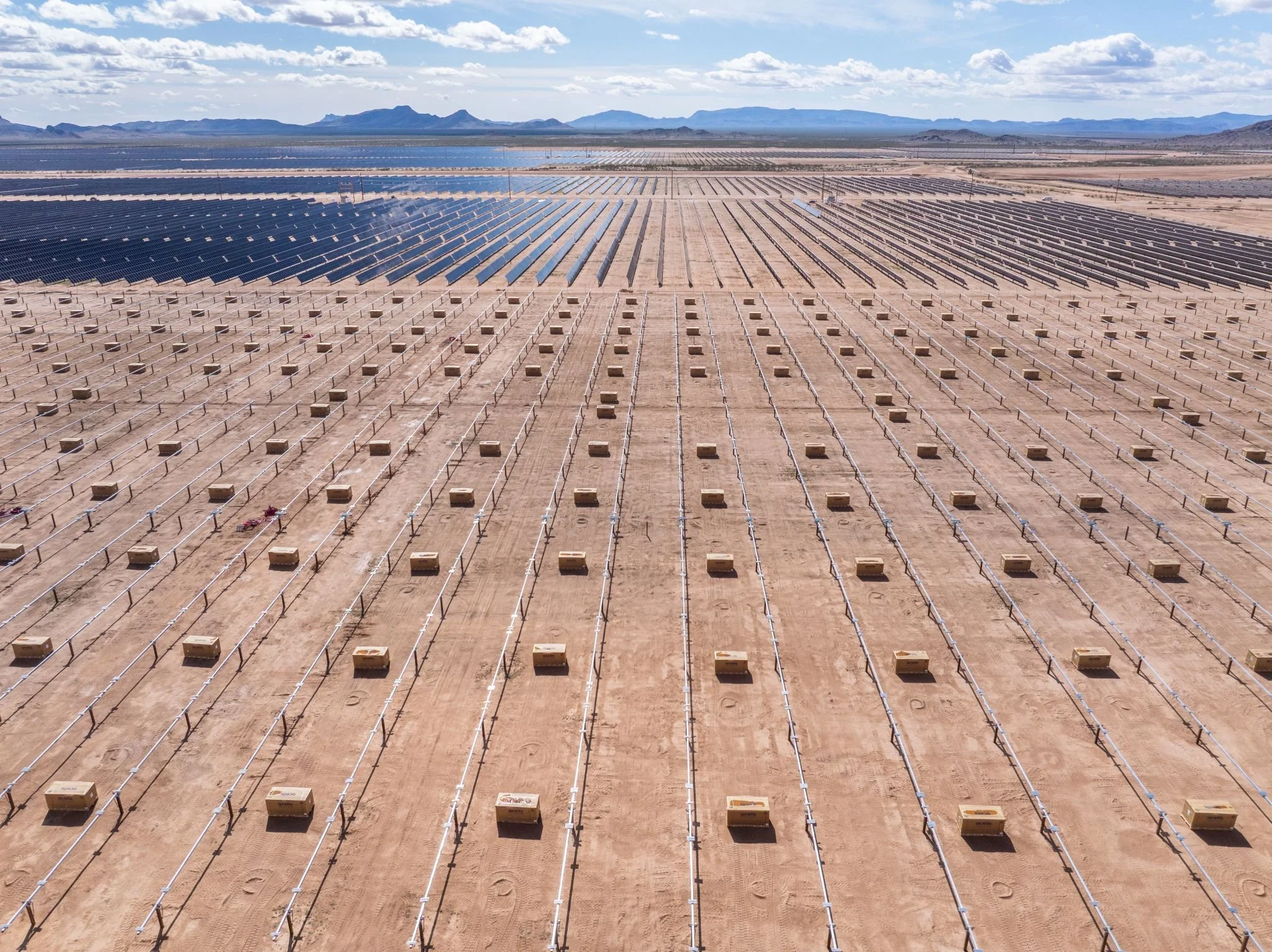 A vast desert landscape with rows of solar panels in the foreground, extending towards distant mountains under a partly cloudy sky.