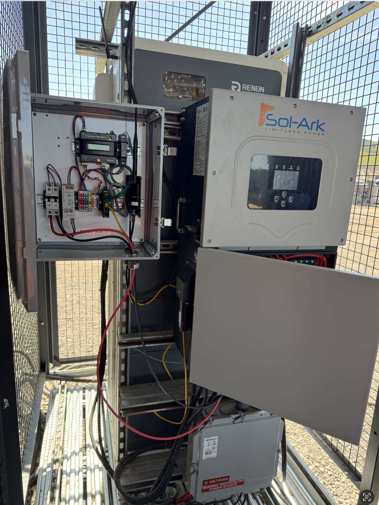 Close-up of electronic equipment inside a metal enclosure, including a control panel, wiring, circuit boards, and a power inverter labeled 'Sol-Ark.' The setup appears to be part of a solar power or energy storage system housed outdoors within a metal framework.