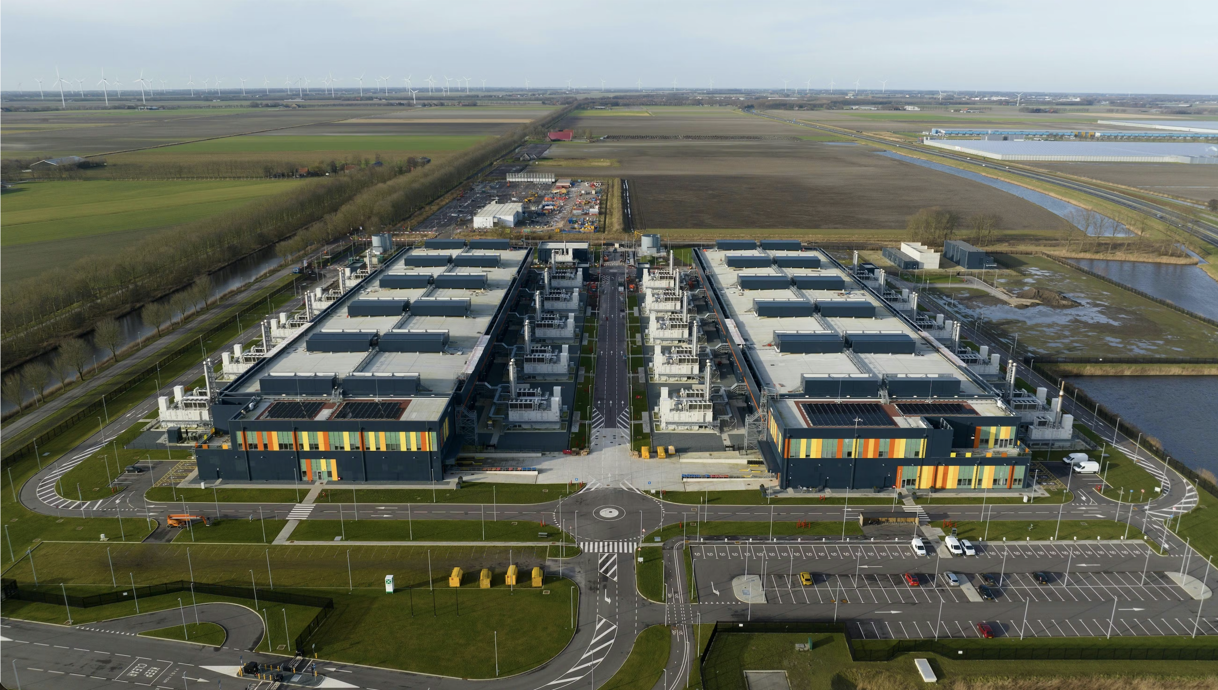 Aerial view of a large modern data center surrounded by roads, parking lots, water channels, and expansive farmland with wind turbines in the distance.