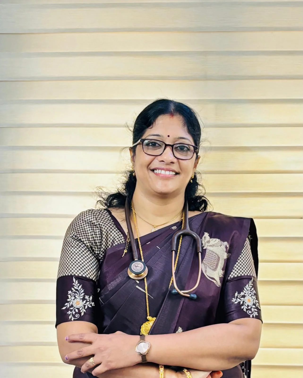 A smiling woman wearing glasses, a traditional saree with intricate embroidery, and a stethoscope around her neck, standing in front of a light-colored, horizontally striped background.