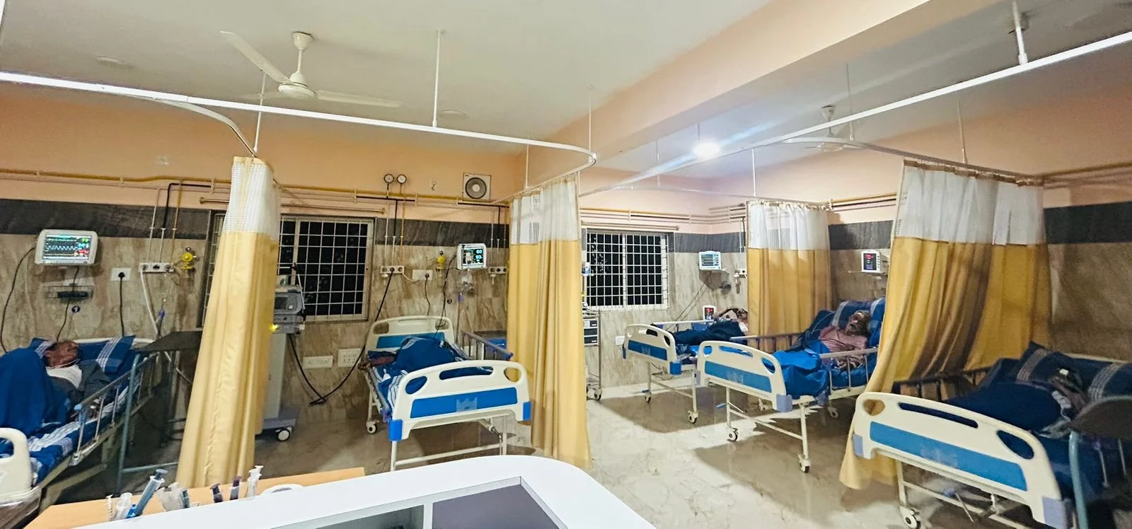 A hospital ward with multiple patients lying in beds, each separated by yellow curtains. Medical monitors are attached to the patients and mounted on the wall, with some connected to medical equipment. The room has beige walls, barred windows, and a ceiling fan.