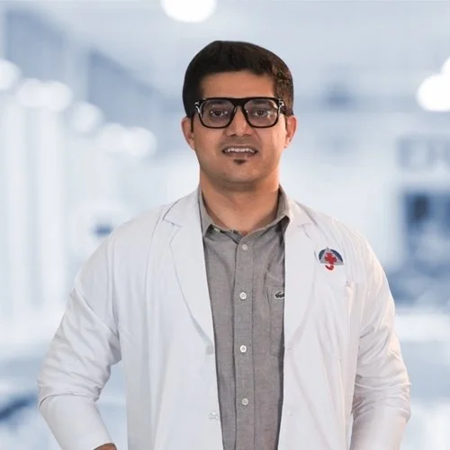 A male doctor wearing glasses and a white lab coat with a medical emblem, standing in a modern clinical setting.