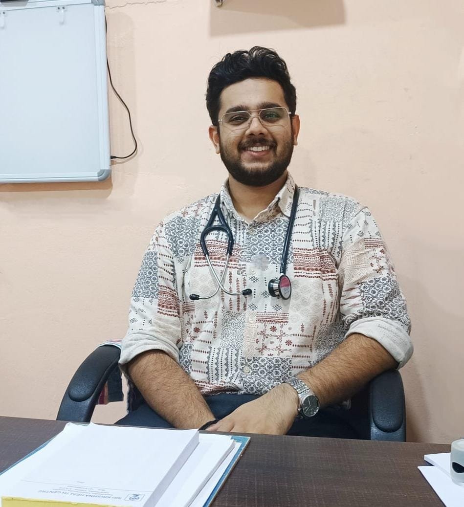 A young male doctor wearing glasses and a patterned shirt with rolled-up sleeves, sitting at a desk with documents, and wearing a stethoscope around his neck, smiling in a medical office.