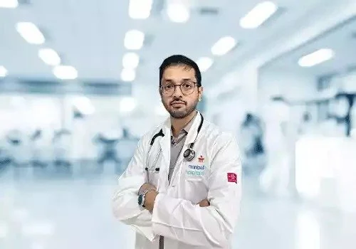A male doctor in a white coat standing in a brightly lit hospital corridor with arms crossed.