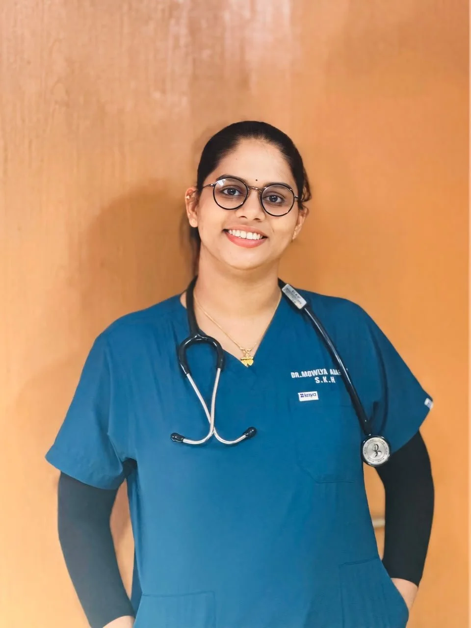 A female healthcare professional wearing blue scrubs and glasses, smiling, with a stethoscope around her neck, standing against a wooden background.