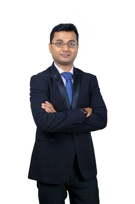 Professional man in a dark suit with arms crossed, wearing glasses and a blue tie, standing against a white background.