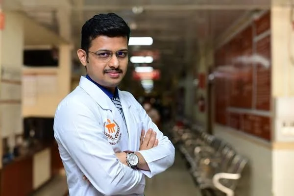 A man with glasses and a mustache wearing a white lab coat standing with crossed arms in a hallway with chairs and walls.