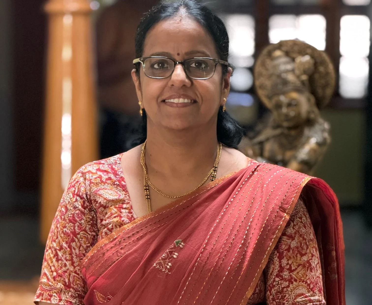 A woman wearing glasses and traditional Indian saree with gold jewelry, smiling indoors with a wooden sculpture in the background.