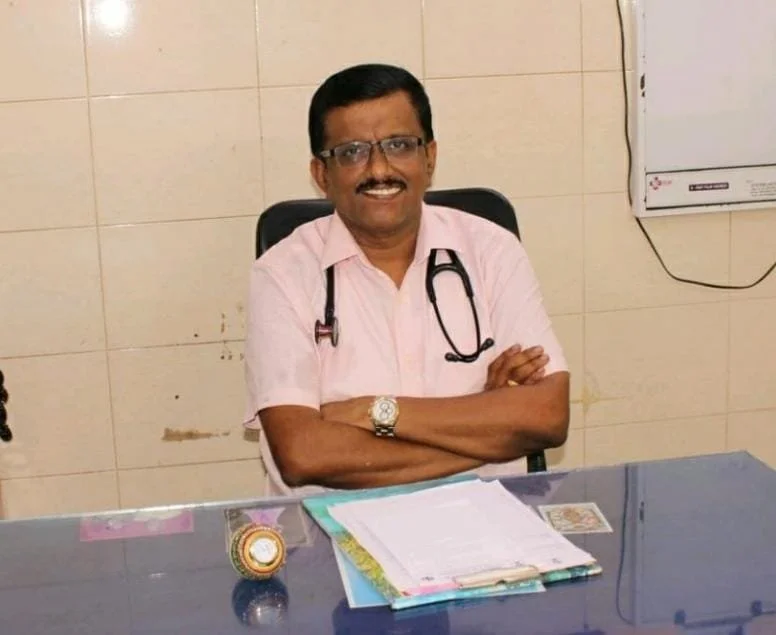 A man wearing glasses and a light pink shirt sitting at a desk with a stethoscope draped around his neck, smiling with arms crossed in a medical office.