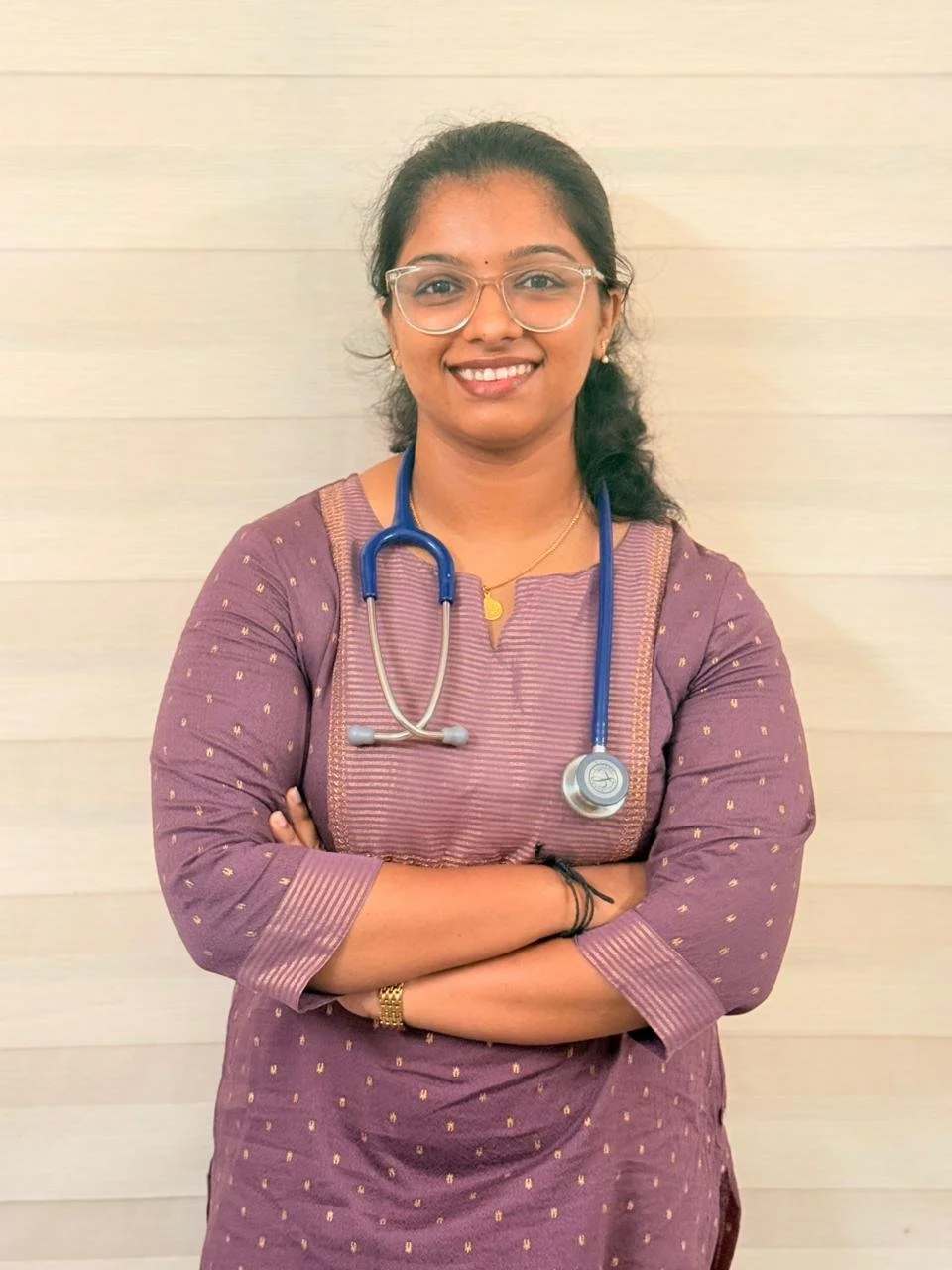 A smiling woman wearing glasses and a purple dress with a stethoscope around her neck, standing against a light-colored wall.