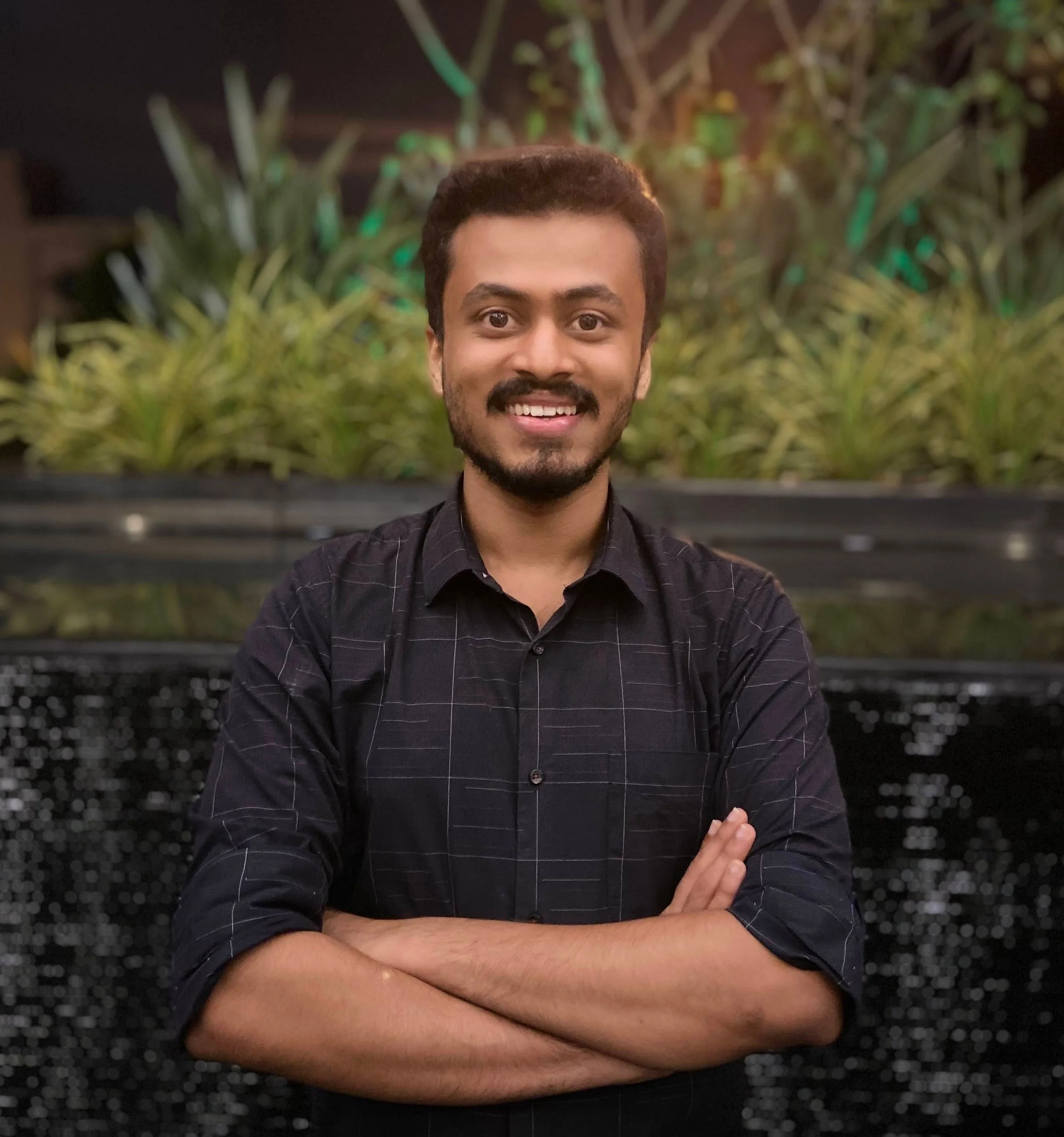A man with dark hair, a beard, and a mustache, smiling with white teeth, crossing his arms, standing in front of green plants and a black and white patterned background at night.
