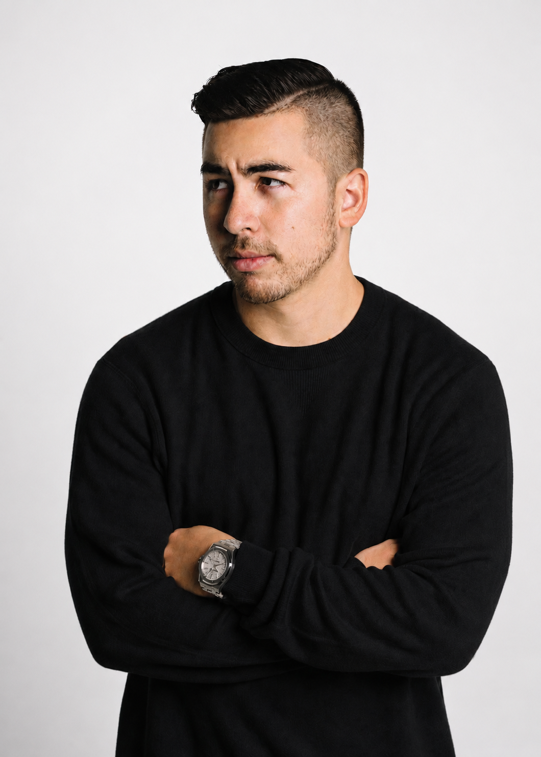 A young man with styled dark hair, wearing a black long-sleeve shirt and a silver watch, looking thoughtfully to the side against a plain white background.