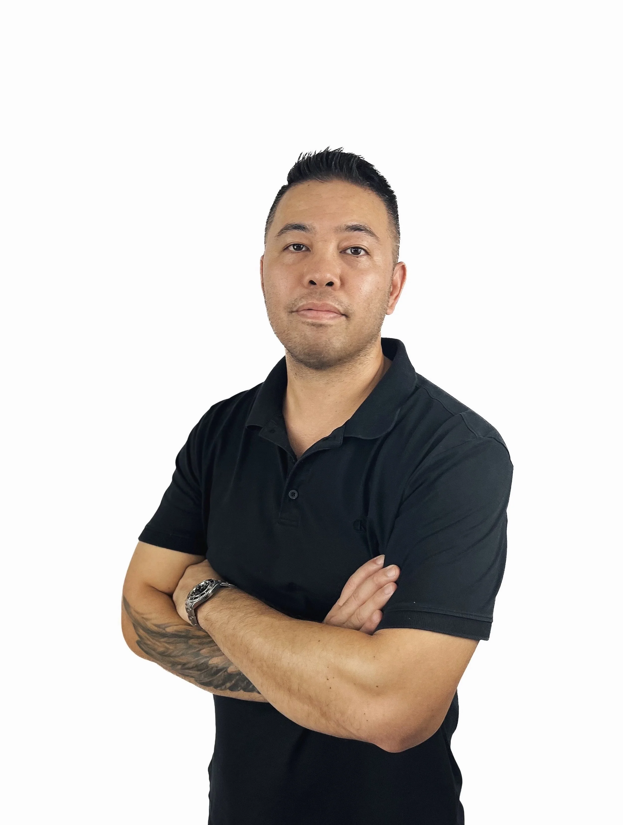 A man with short black hair and tattoos on his left arm, wearing a black polo shirt, standing with arms crossed against a white background.