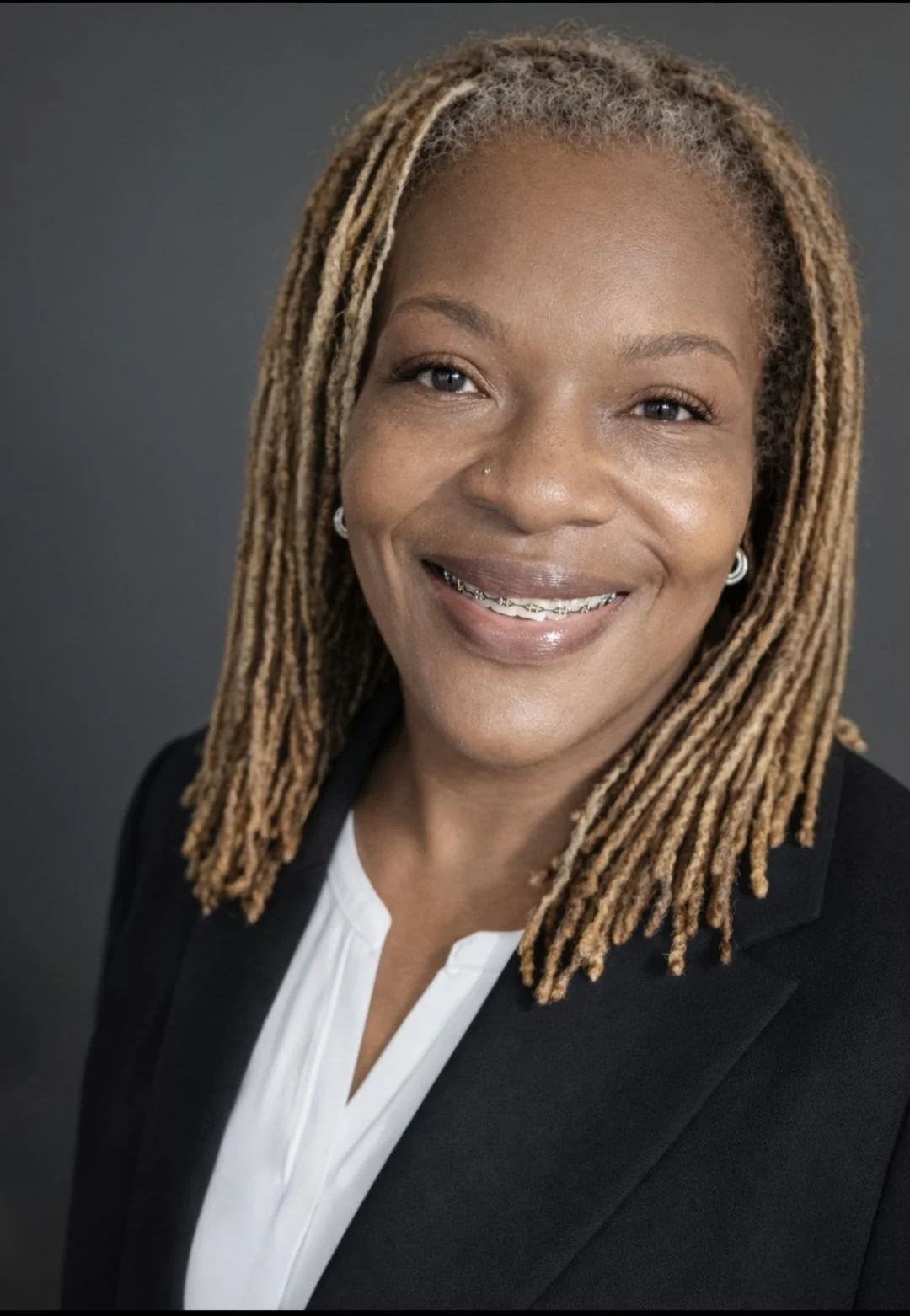 Professional headshot of a smiling Black woman with blonde dreadlocks, wearing a black blazer and white blouse, against a dark gray background.
