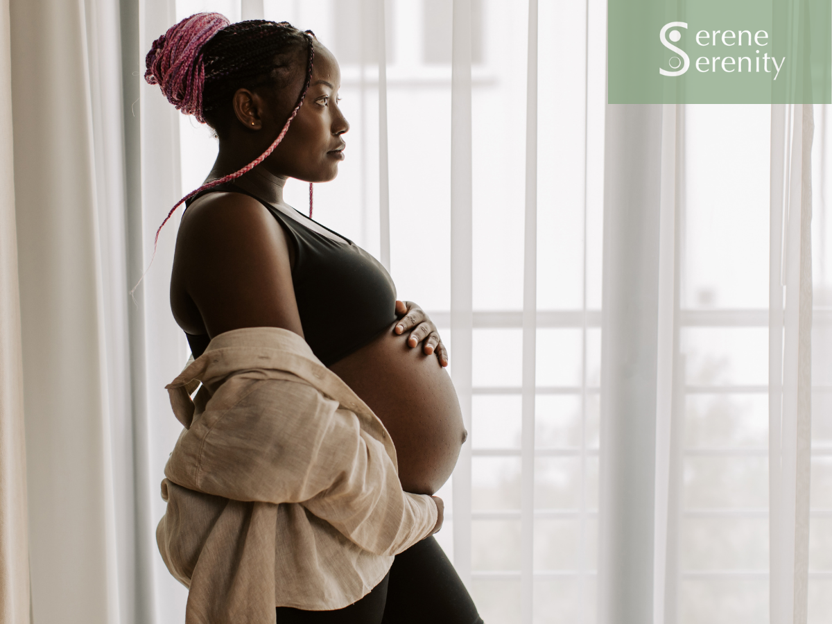 Pregnant woman with pink braided hair standing by window with sheer curtains, wearing black sports bra and beige shirt draped over shoulders, looking contemplative. fertility support