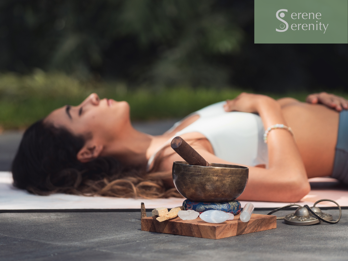 A woman lying on her back on a yoga mat outdoors with eyes closed, surrounded by crystals, a singing bowl, and a bell, with a background of greenery. Nervous system regulation