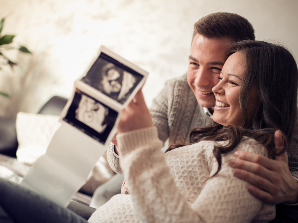 A couple holding ultrasound images, smiling, with a woman in the foreground, and the man and woman sitting closely together