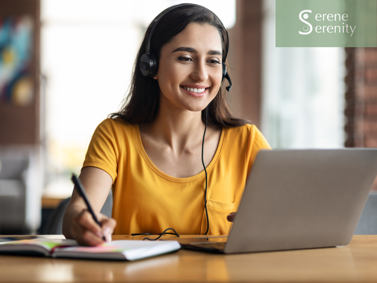 A woman wearing a yellow shirt and headset smiling while working on a laptop at a desk, with a notebook open and a pen in her hand.