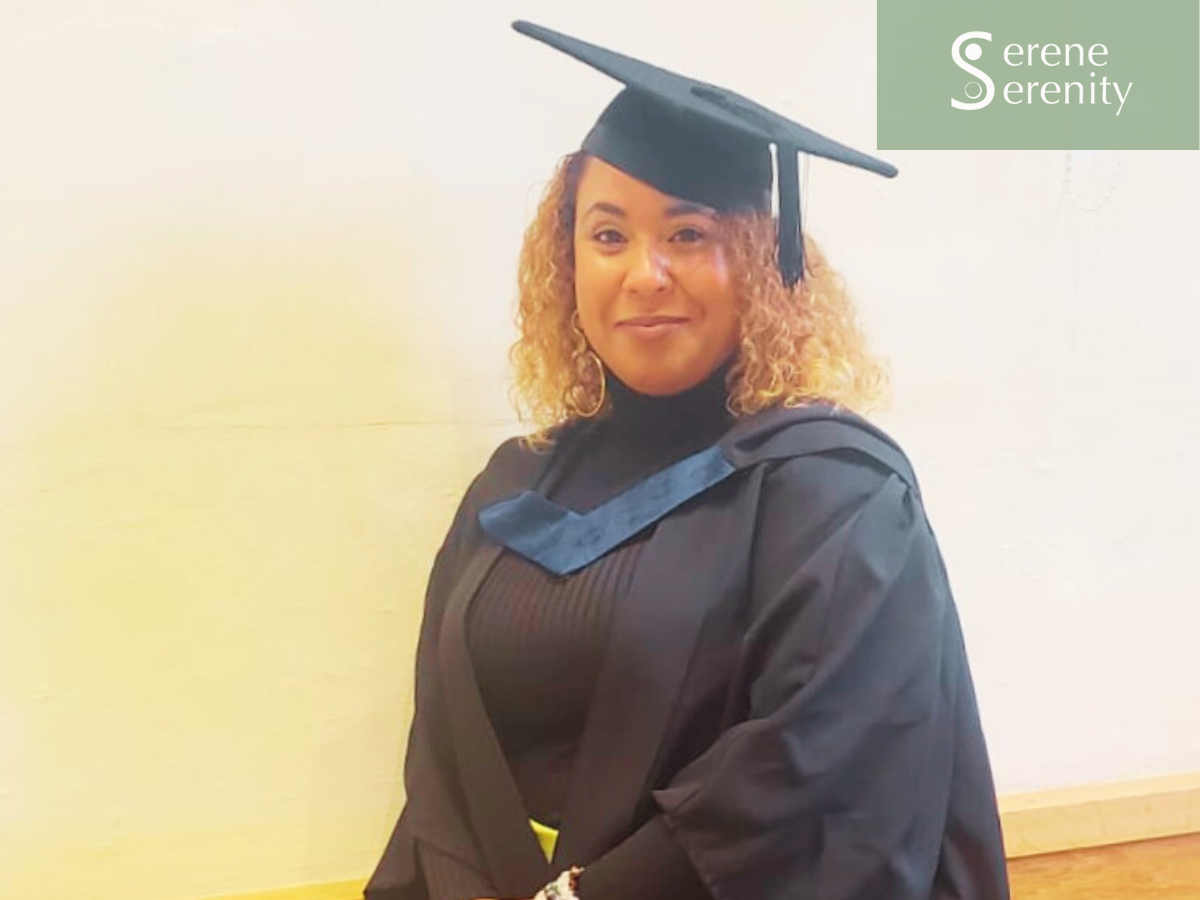 A woman in a graduation cap and gown smiling, posing indoors with a beige wall in the background.