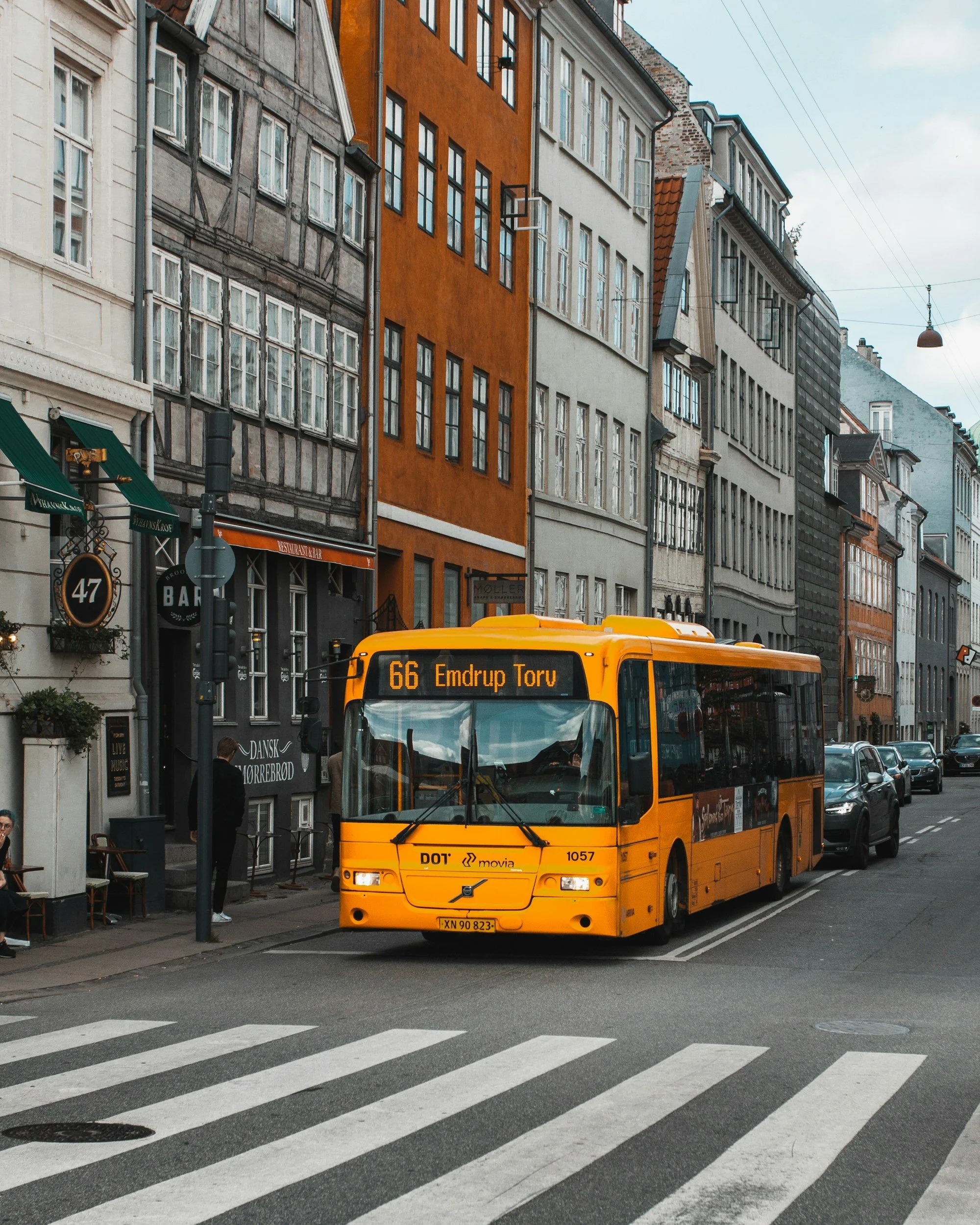A city street with a yellow bus in the foreground, showing route number 66 to Emdrup Torv. The street has a pedestrian crossing and parked cars along the side. Buildings with various architectural styles and colors line the street, including a notable orange-colored building. People are walking near street cafes and shops.