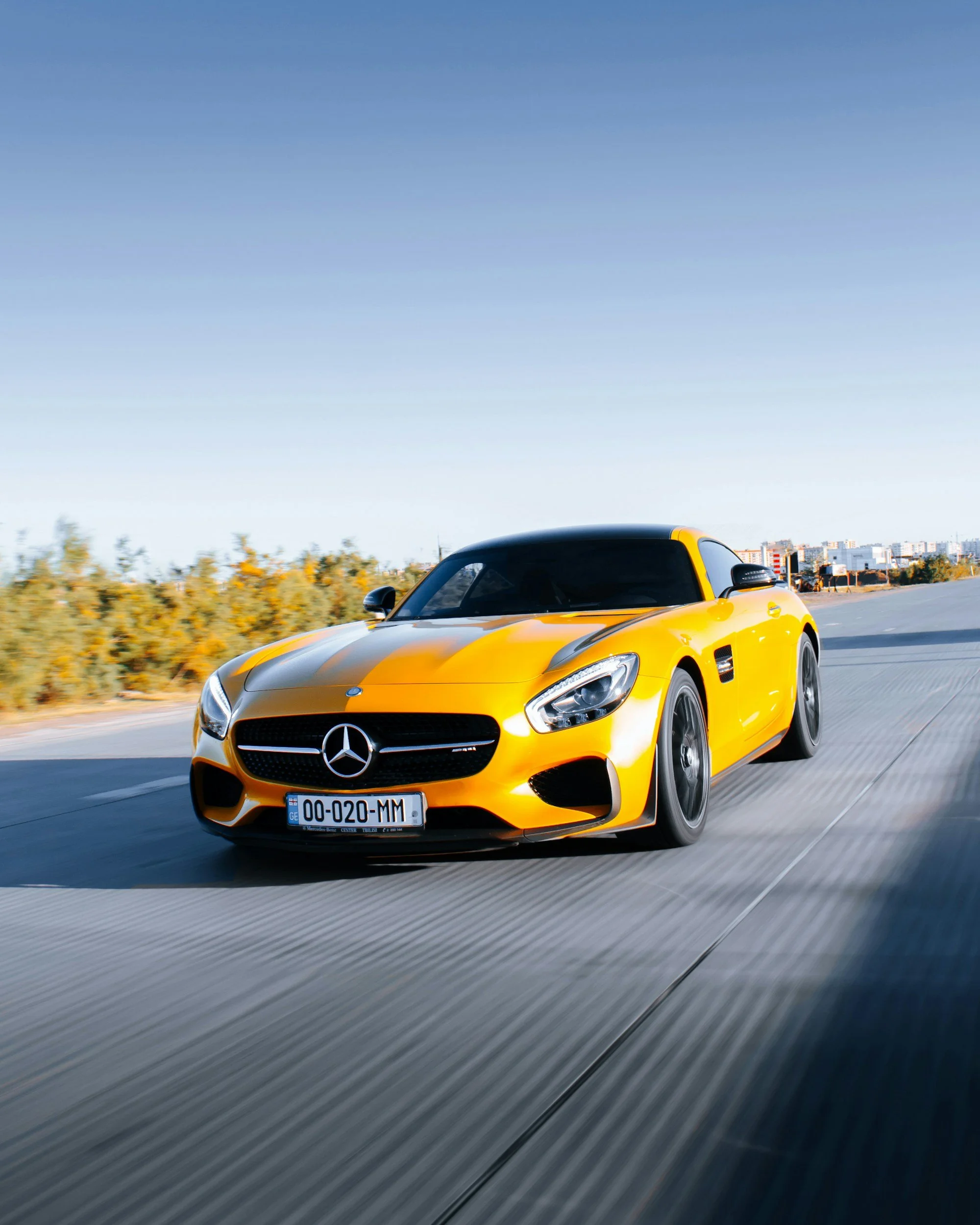A yellow Mercedes-Benz sports car driving on a highway with blurred background of trees and buildings, under a clear blue sky.