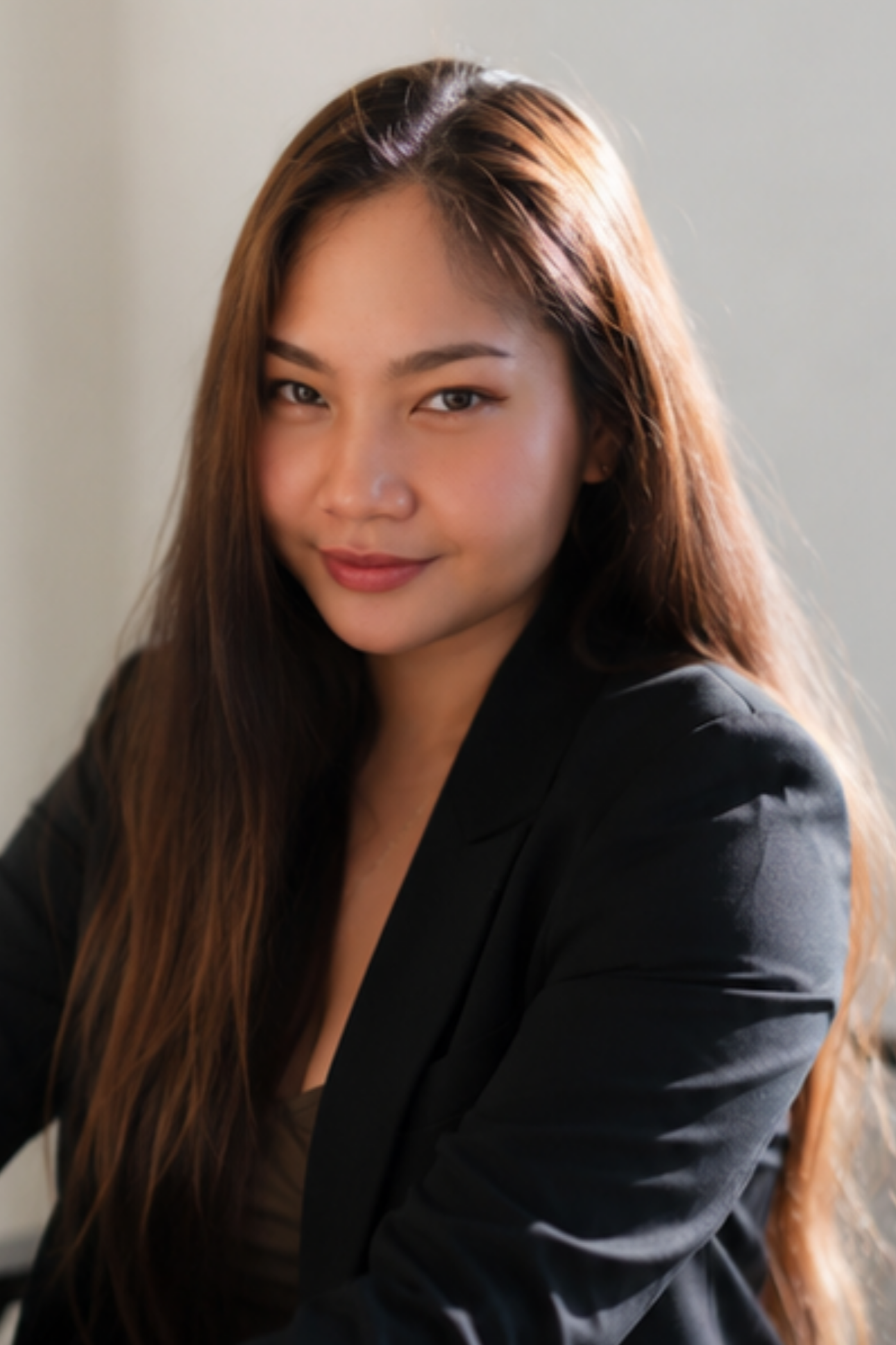 Close-up of a young woman with long brown hair, wearing a black blazer, smiling softly, against a plain background.