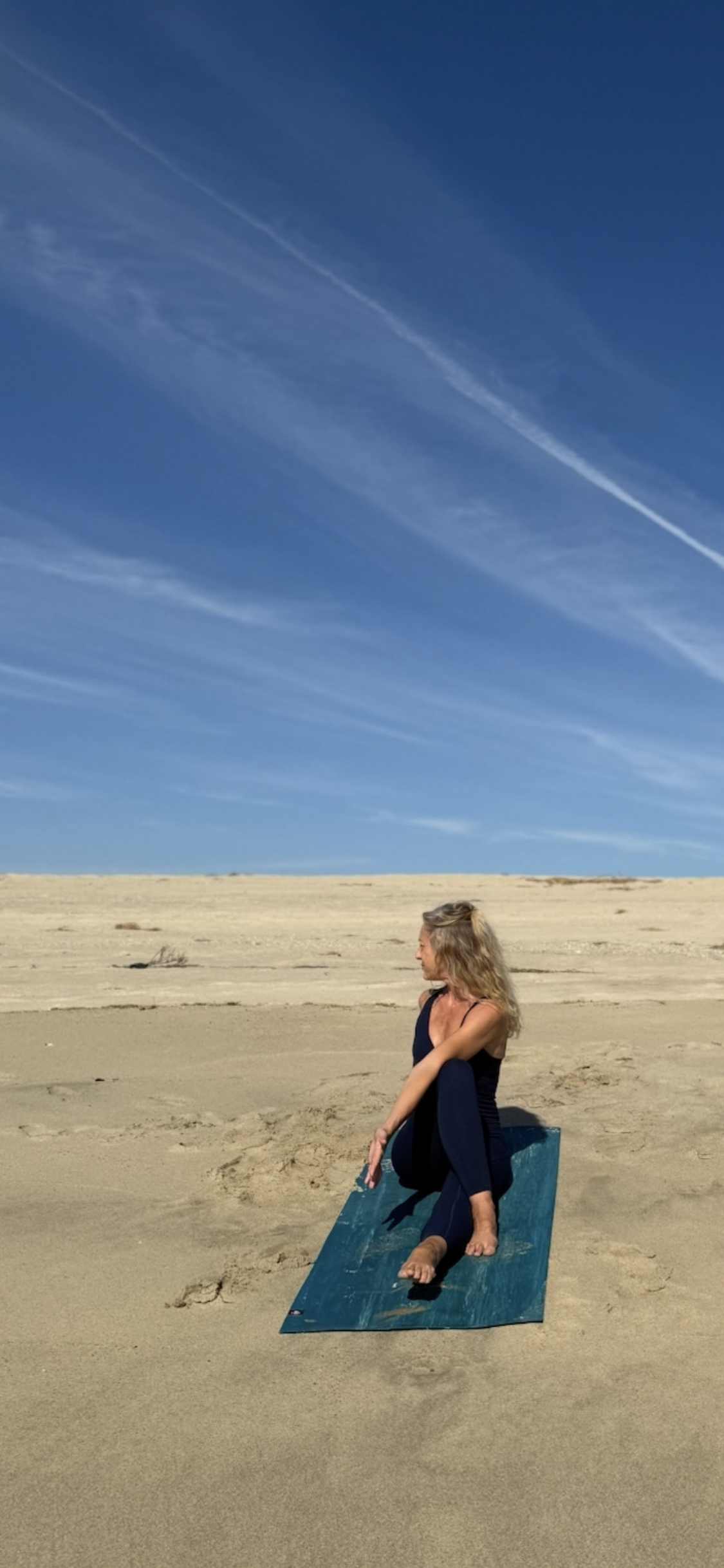 A woman practicing yoga on a blue mat on a sandy beach under a blue sky with wispy clouds.