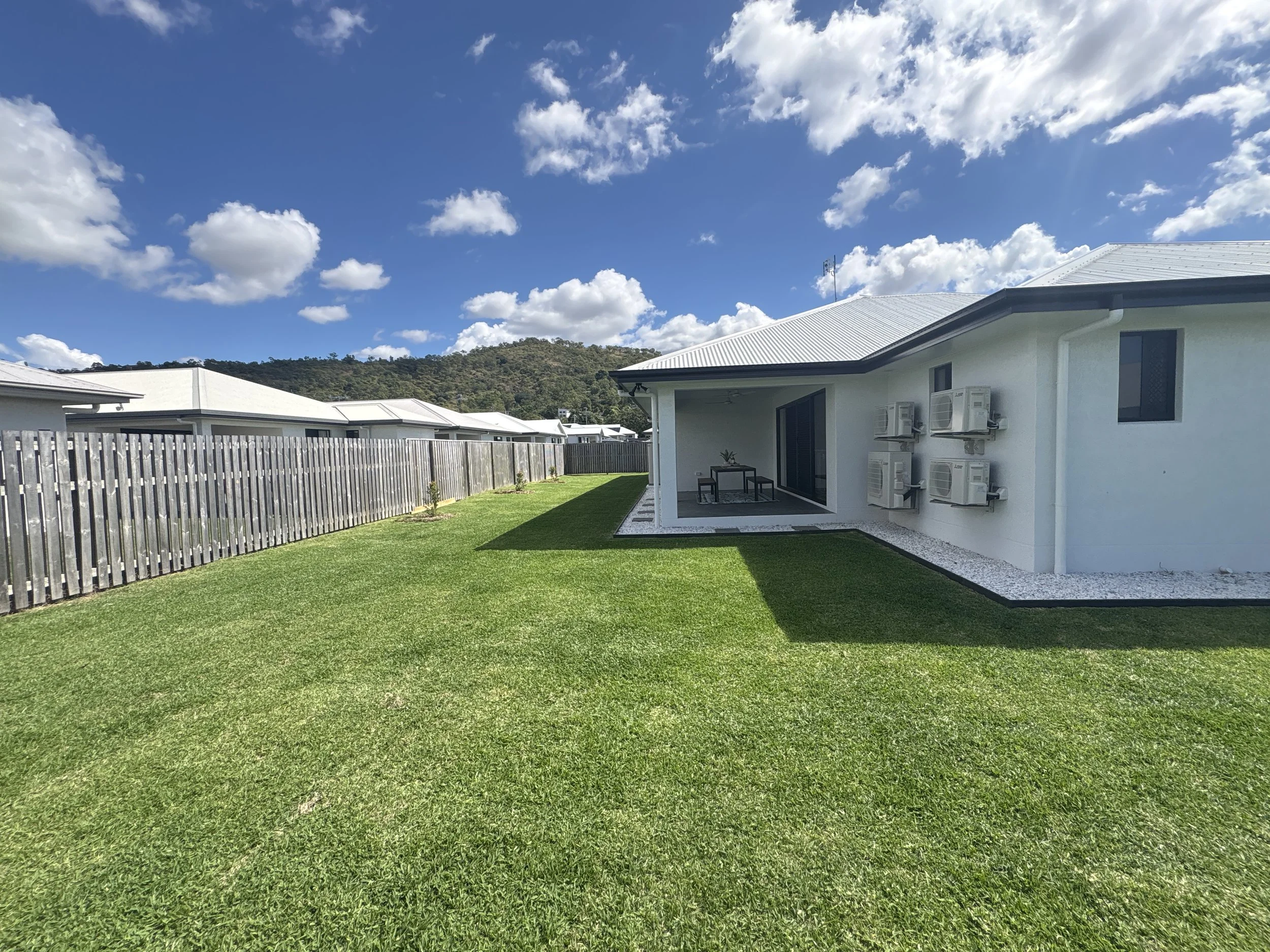 A backyard with well-maintained green grass, a white house with air conditioning units on the exterior wall, a fencing on the left, and a mountain with trees in the background under a partly cloudy blue sky.