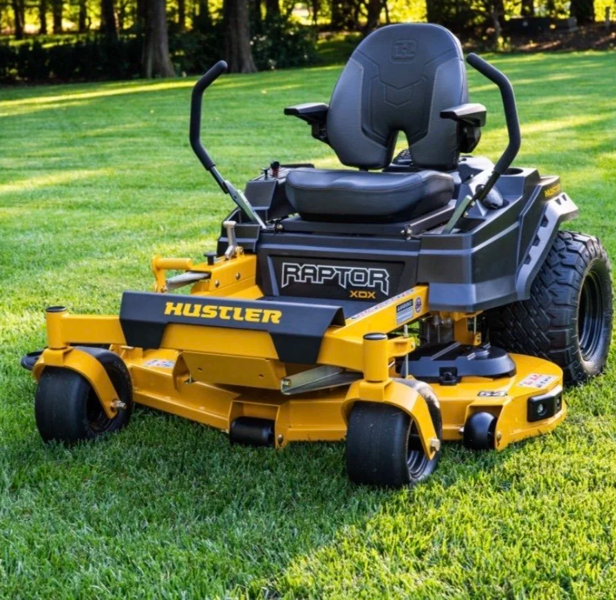 Yellow Hustler Raptor riding lawnmower on a green grassy field with trees in the background.