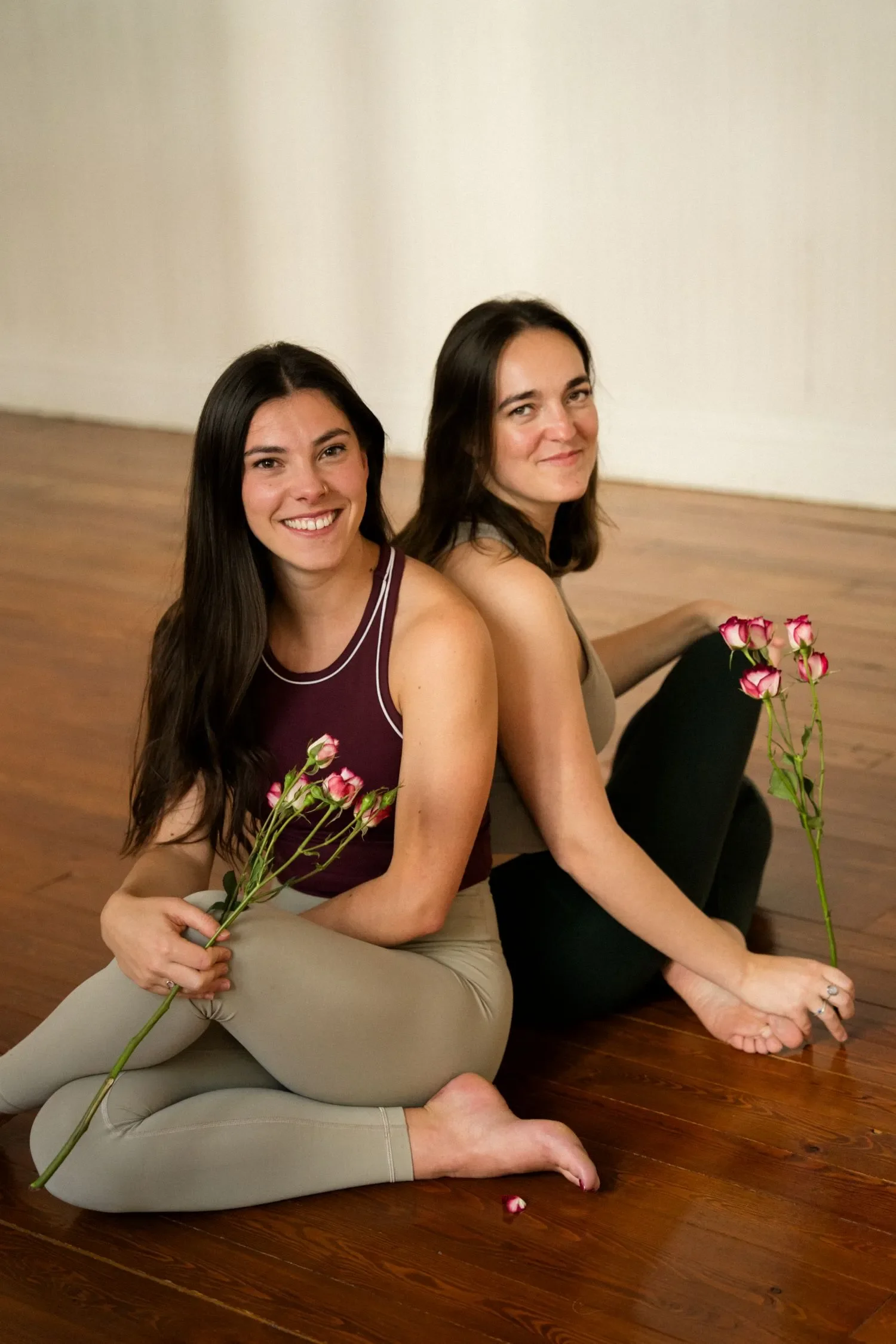 Dos mujeres sentadas en el piso de madera, una con camiseta rosa y la otra con camiseta gris, ambas sosteniendo flores rosadas y sonriendo.
