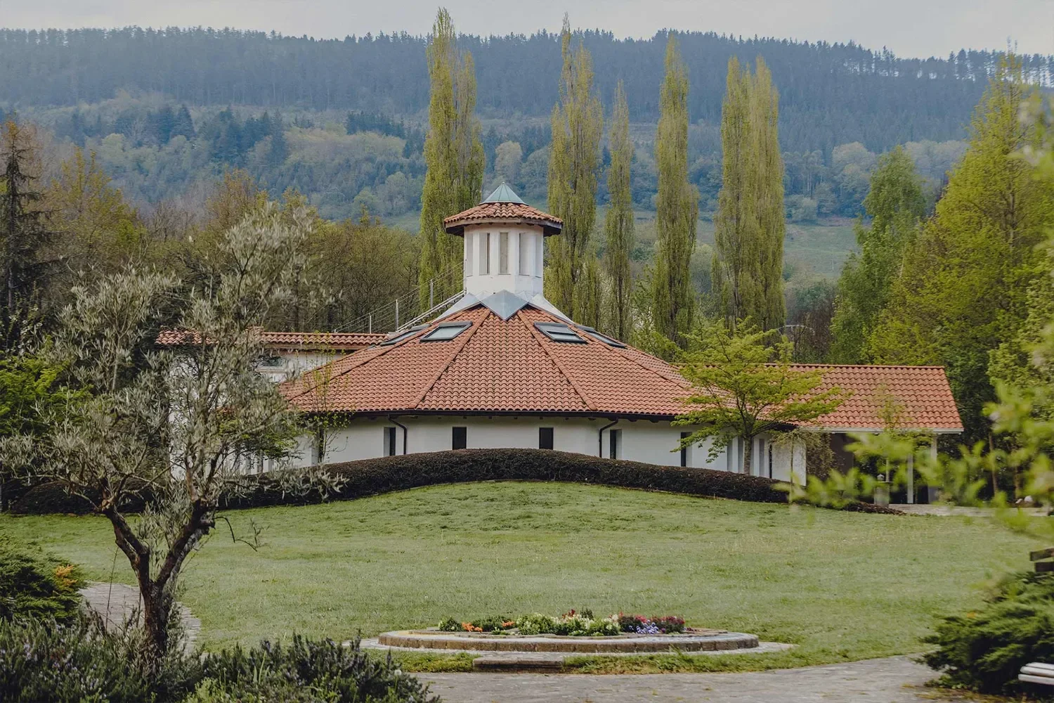 Casa con tejado de tejas rojas, en un entorno natural con árboles altos y colinas al fondo, en un día nublado.