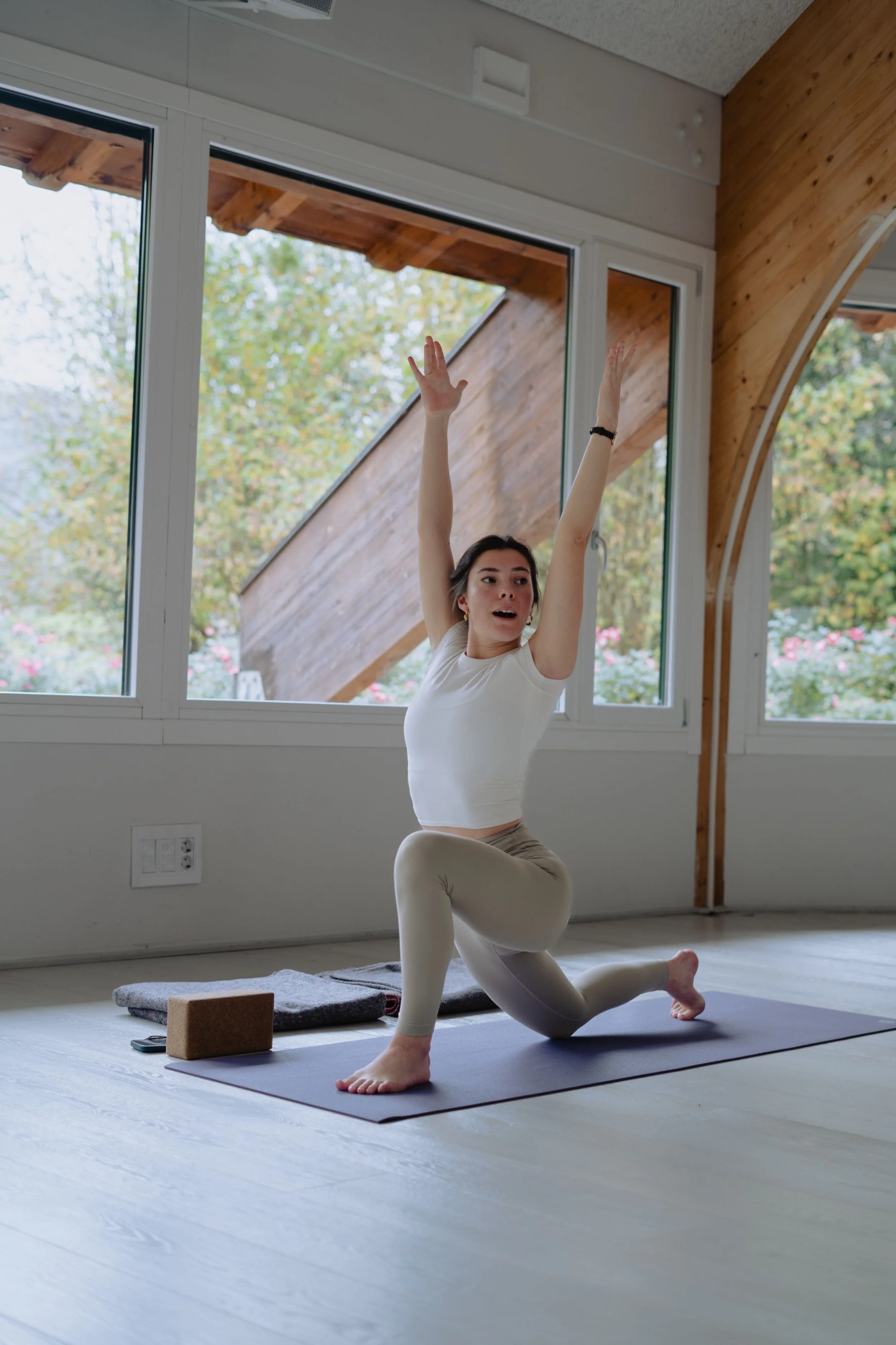 Mujer practicando yoga en interior con ventanas grandes y vista a la naturaleza, usando ropa blanca y en posición de equilibrio.