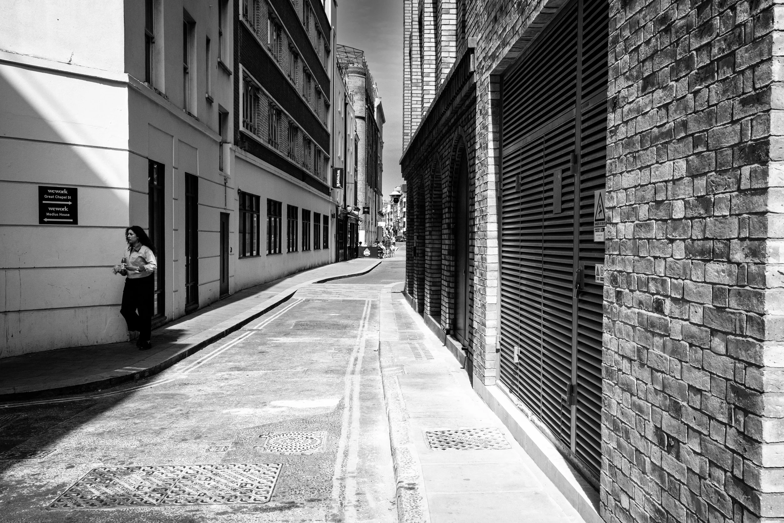 A black and white photo of a narrow urban street with tall buildings on both sides. A woman is walking on the sidewalk on the left, holding a cup. The street has a slight curve, and there are placards and vent covers on the pavement, with some signage visible on the buildings.