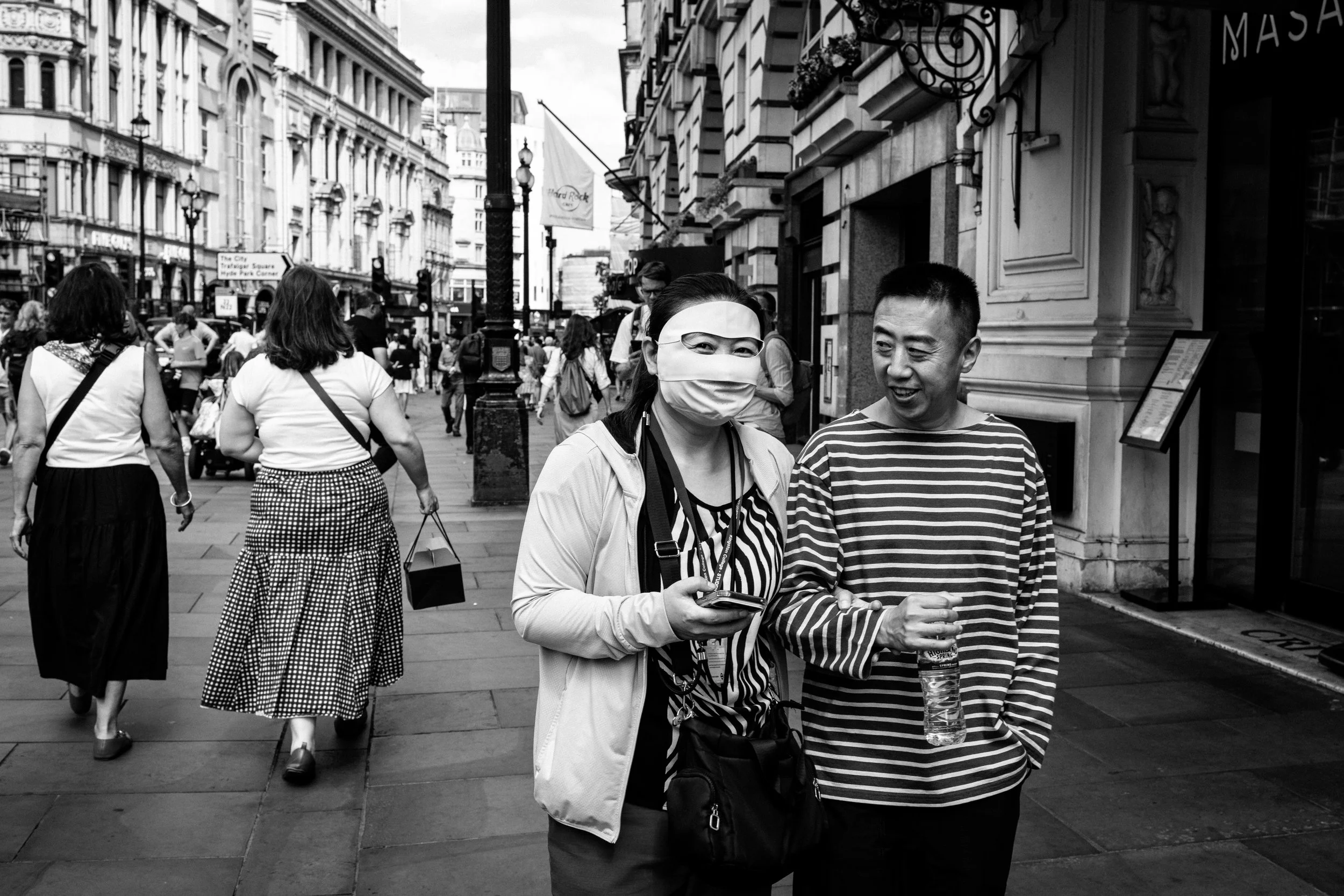A busy city street scene in black and white with diverse pedestrians walking. Two people in the foreground are smiling, one with a mask, holding a phone and a bottle of water. Other pedestrians are walking in the background, along with buildings and street signs.