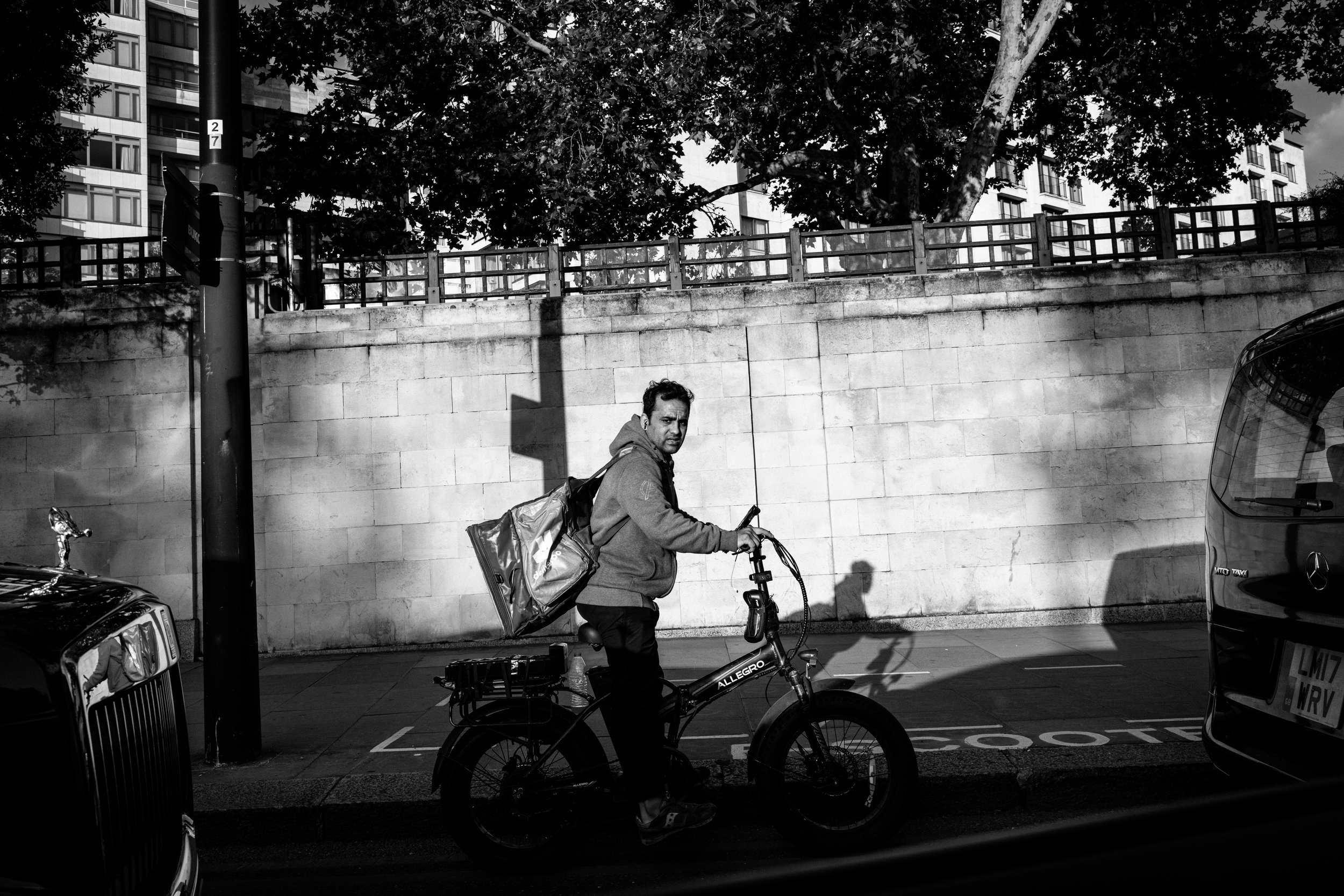 A man riding a bicycle on a city street, carrying a large backpack, with cars parked along the sidewalk and a stone wall with trees above it in the background, in black and white.