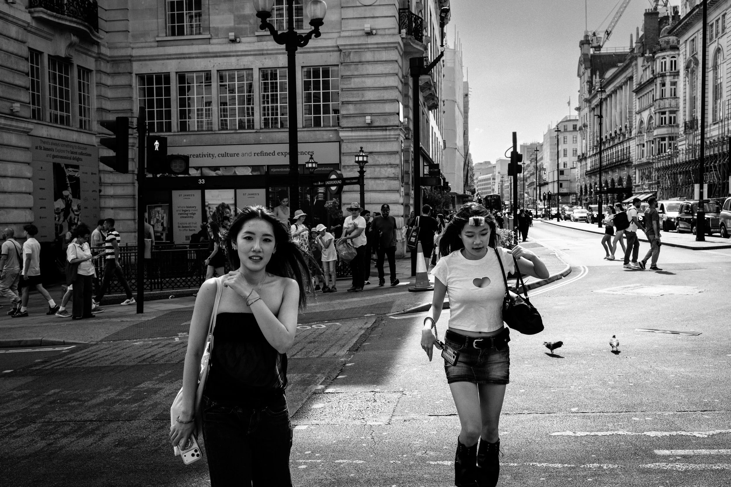 Two young women walking across a city street in London, with a crowd and historic buildings in the background.