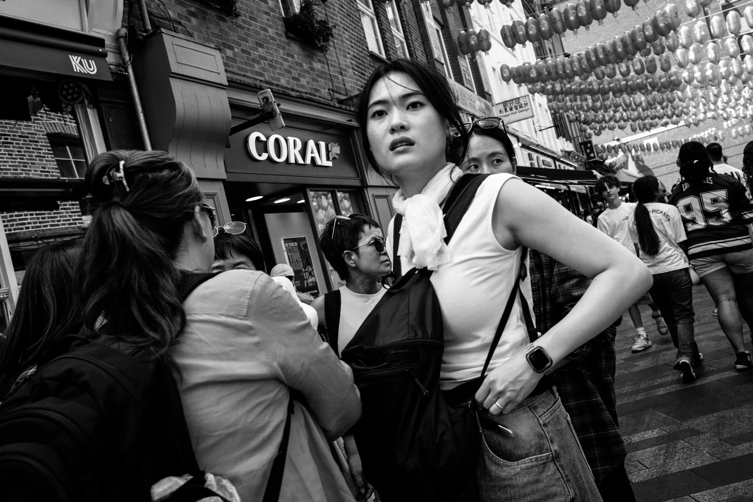 A black-and-white photo of a crowded street with hanging lanterns and various pedestrians, including a young woman with short dark hair, a scarf around her neck, sunglasses on her head, and a serious expression, looking to the side.