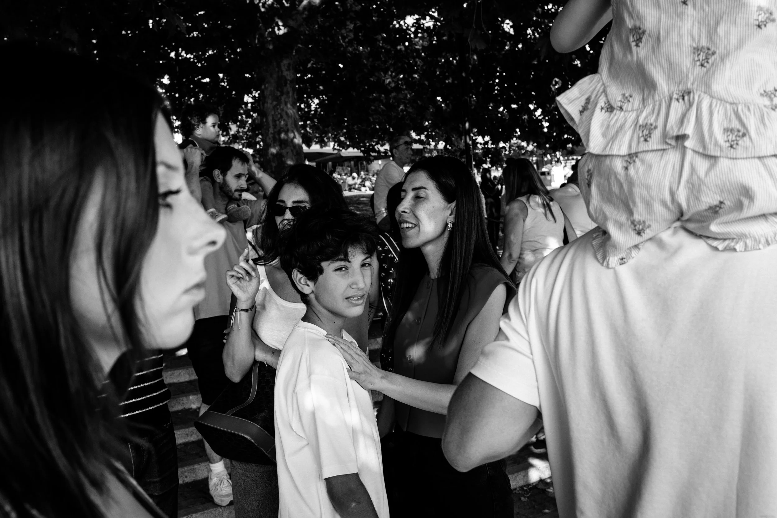 People gathering outdoors under trees, engaging in conversation, with some standing and some sitting.