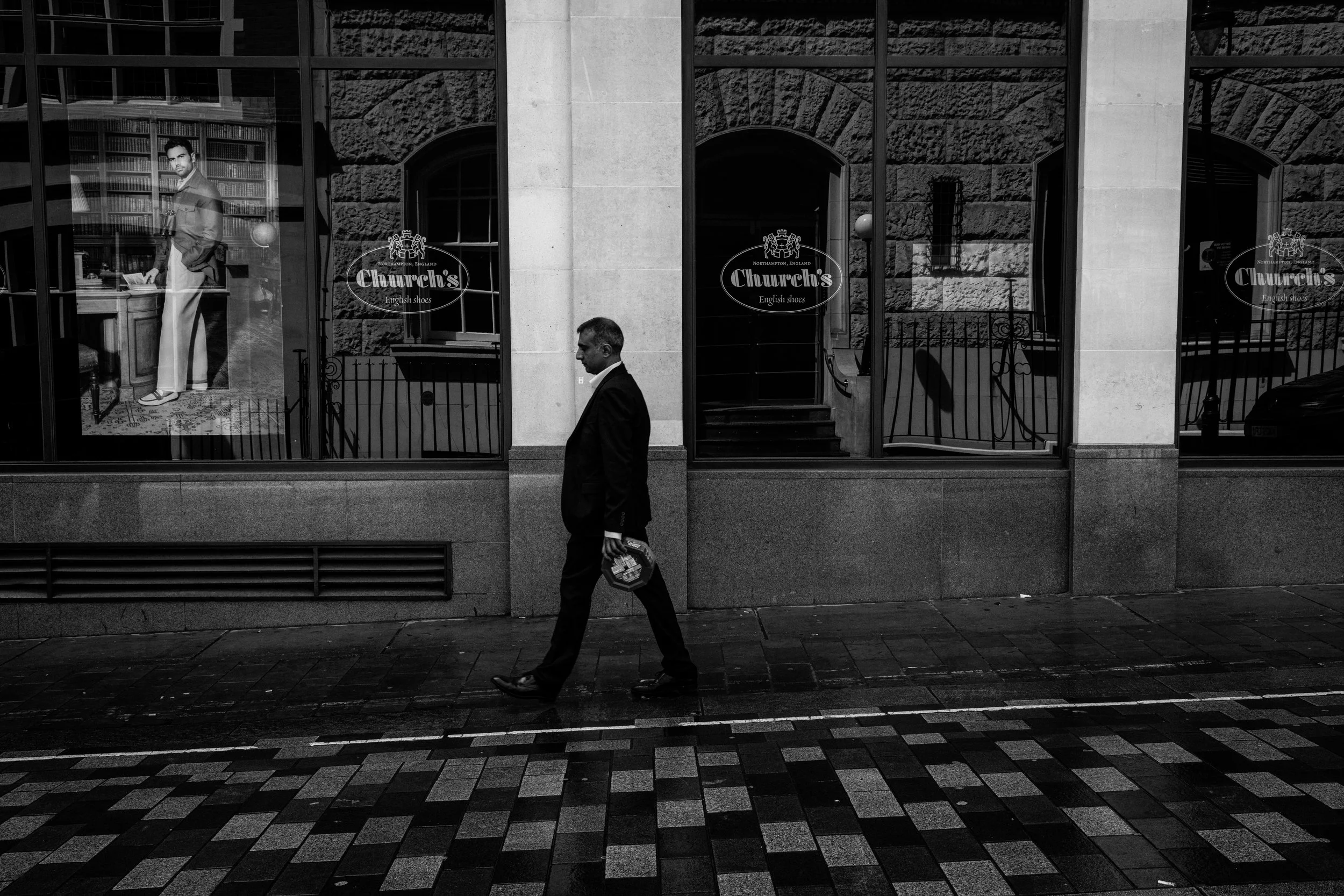 A man in a dark suit walking past a storefront with signs for a shoe store called 'Churchy's' in Northampton, England. The storefront has large windows showing mannequins and interior furniture, with a second man visible inside the store.